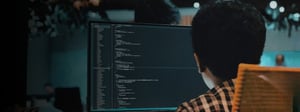 A software developer works at a desktop computer displaying code on a large monitor in a dimly lit workspace, representing cloud database development, programming, and modern IT operations.