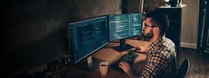A cybersecurity analyst working at a desk with dual monitors displaying code, representing SOC outsourcing and managed security operations.