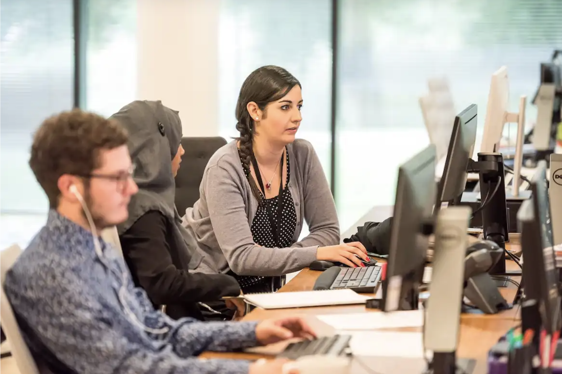 Healthcare support staff work at computer stations in a shared office, monitoring systems and collaborating in a technology-enabled service desk environment supporting healthcare operations.